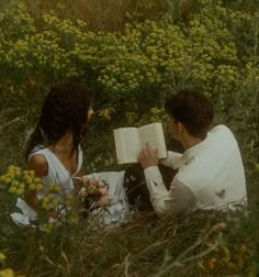 a man and woman are sitting in the grass reading a book while looking at each other