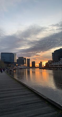 people are walking on the boardwalk near the water and buildings in the background at sunset