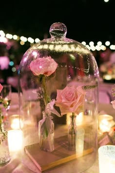 a glass clochet with pink roses in it sitting on a table next to candles