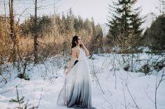 a woman in a long dress standing in the snow with trees and bushes behind her