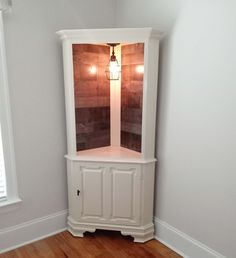 a corner cabinet in the corner of a room with wood floors and white walls, along with a wooden floor