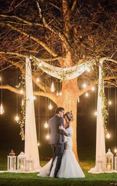 a bride and groom standing in front of a tree with lights strung from the branches