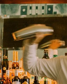 a man in a white coat is making a drink at a bar with liquor bottles behind him