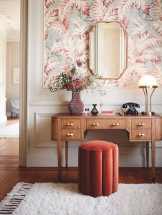 a desk with a mirror, stool and flowers on it in front of a wall