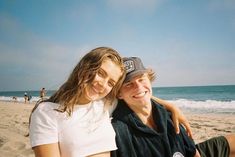 a man and woman sitting on the beach with their arms around each other, smiling at the camera