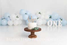 a white cake sitting on top of a wooden stand next to blue and white balloons