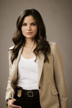 a beautiful young woman wearing a tan blazer and black pants standing in front of a gray background