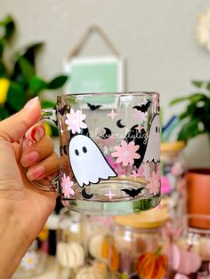 a hand holding a glass cup with a ghost on it and flowers in the background