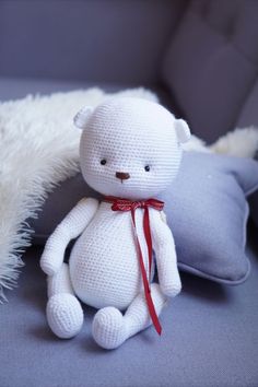 a white crocheted teddy bear sitting next to a pillow on a couch with a red ribbon around its neck