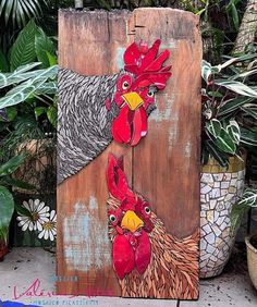 two roosters painted on wooden boards in front of potted plants and greenery