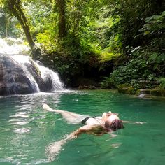 a woman is floating in the water near a waterfall and some green trees with leaves on it