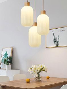 three lights hanging over a wooden table with white chairs and flowers in a vase on it