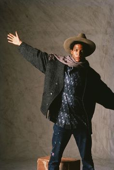 a man in a hat and jacket is standing on a suitcase with his arms outstretched