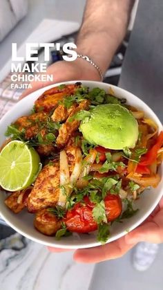 a person holding a white bowl filled with chicken, tomatoes and lettuce next to a lime