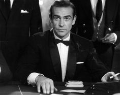 a man in a tuxedo sitting at a desk with other men behind him