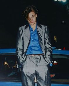 a man standing next to a blue car wearing a gray suit and blue shirt with his hands in his pockets