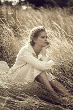 a woman is sitting in tall grass with her hand on her chin and looking off into the distance