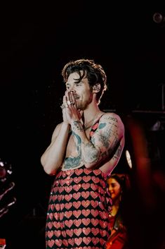 a man with tattoos on his chest standing in front of a microphone and holding his hands to his mouth