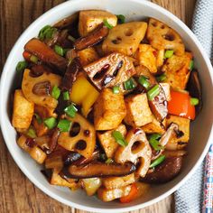 a white bowl filled with tofu and vegetables on top of a wooden table next to a blue napkin