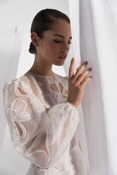 a woman wearing a sheer white dress and holding her hand up to the side while standing in front of a curtain