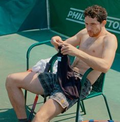 a shirtless man sitting in a chair on a tennis court