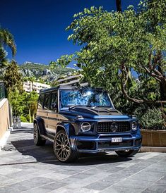 a blue mercedes g - class is parked in front of some trees and bushes on a sunny day