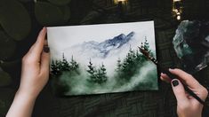 a person holding a paintbrush and painting a landscape with watercolors on paper