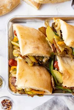 two trays filled with sandwiches on top of a white countertop next to bowls of seasoning