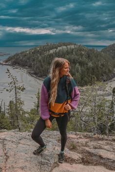 a woman standing on top of a rock next to the ocean and trees in the background