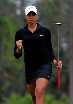 a woman in black shirt and skirt holding a red golf club while walking across a green field