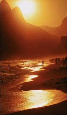 people are walking on the beach as the sun goes down in the mountains behind them