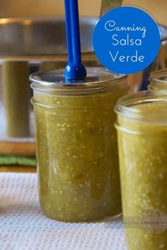 two jars filled with green salsa sitting on top of a white towel next to a blue spoon