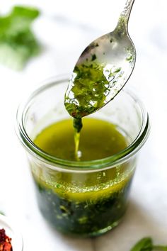 a spoon full of green liquid being poured into a small glass jar filled with greens