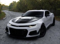 a white camaro with black stripes is parked on the side of a dirt road