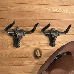 two metal steer head hooks on a wooden wall next to a hat and leather bag