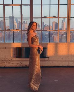 a woman in a gold dress standing next to a window looking out at the city