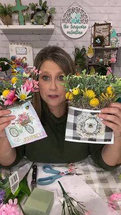 a woman sitting at a table with flowers and cards in front of her, holding up two