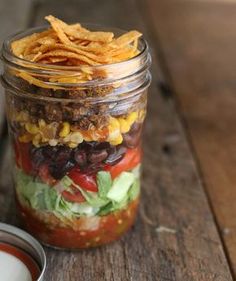 a mason jar filled with taco salad next to a container of ranch dressing on a wooden table