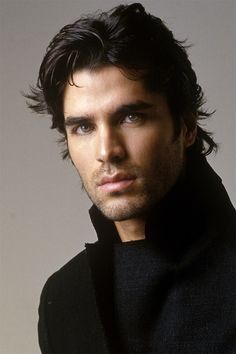 a man with long hair wearing a black coat and looking at the camera while standing in front of a gray background