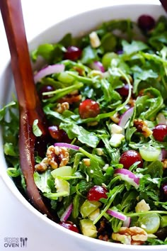 a salad in a white bowl with wooden utensils on the side, topped with cranberries and walnuts