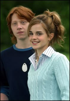 two young people standing next to each other in front of green trees and bushes, one wearing a blue sweater