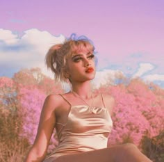 a woman sitting on top of a rock in front of trees and pink flowers with her legs spread out