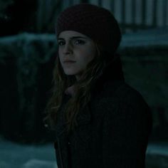 a woman with long hair wearing a black coat and a red beanie standing in the dark