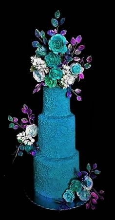 a three tiered blue cake with flowers and leaves on the top, against a black background