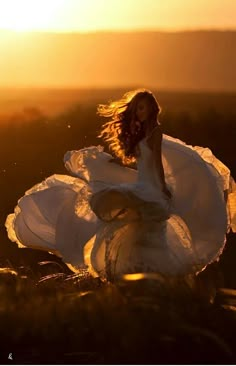 a woman in a white dress sitting on top of a grass covered hill at sunset