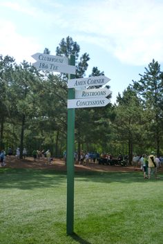 a sign post in the middle of a grassy area with people walking around and trees behind it