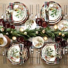 the table is set for christmas dinner with plates, silverware and greenery on it