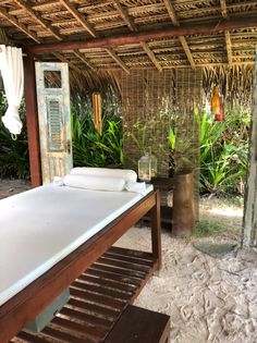 a bed sitting under a thatched roof next to a lush green field and trees