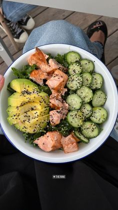 a person holding a white bowl filled with salmon, cucumbers and avocado