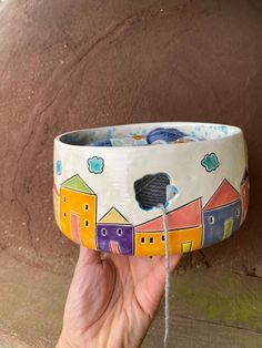 a hand holding a painted bowl in front of a brown wall with houses on it